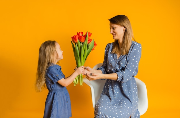 La pequeña hija le da a su madre un ramo de tulipanes en una pared amarilla