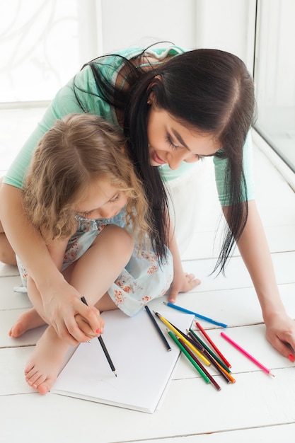 Pequeña hija linda y su madre dibujando con lápices de colores y divirtiéndose juntos. Niño bonito y mamá jugando en el interior. Familia feliz pasar tiempo dibujando.