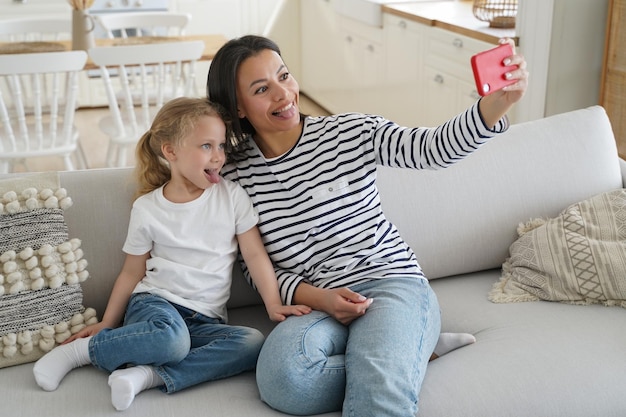 La pequeña hija madre muestra lenguas tomando selfie de la familia con el teléfono divirtiéndose juntos en casa