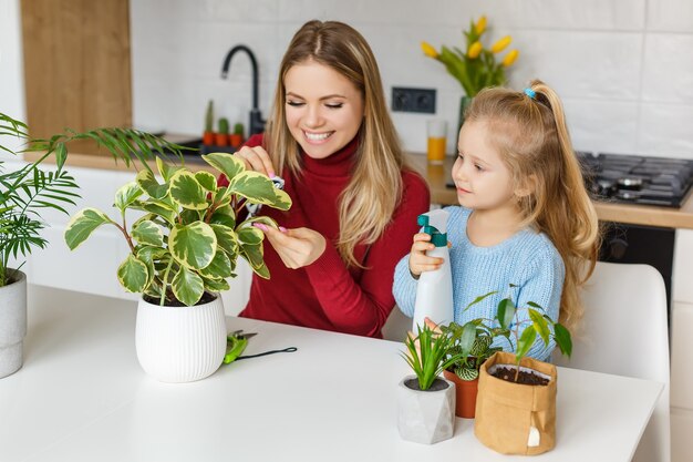 Pequeña hija y madre rociando y limpiando plantas de interior. Niño ayudando a mamá a cuidar las plantas