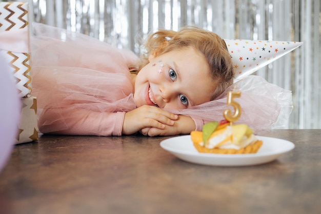 Pequeña y linda cumpleañera posando cerca de la torta de cumpleaños en una hermosa foto horizontal de vestido festivo rosa