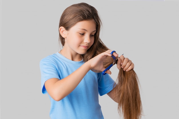 Pequena linda garota de camiseta azul, olhando para os cabelos longos naturais e vai cortá-lo com uma tesoura azul