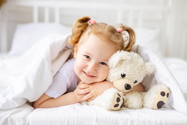 Una pequeña y linda niña se asoma por debajo de una manta en una cama de algodón blanco en casa sonriendo abrazando a un oso de peluche