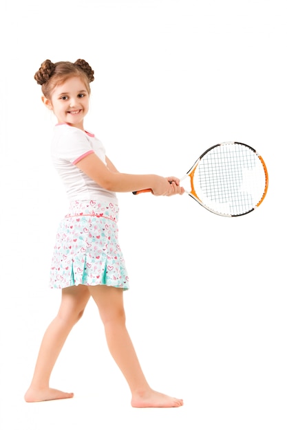 Pequena menina positiva em roupas elegantes em pé e segurando a raquete de tênis na mão e sorrindo sobre fundo branco