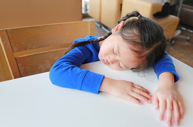 Pequeña muchacha asiática cansada del niño que duerme en la tabla blanca en el café.