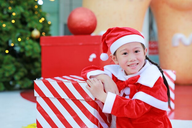 Pequeña muchacha asiática linda feliz del niño en el traje de santa con la caja de regalo cerca del árbol de navidad y
