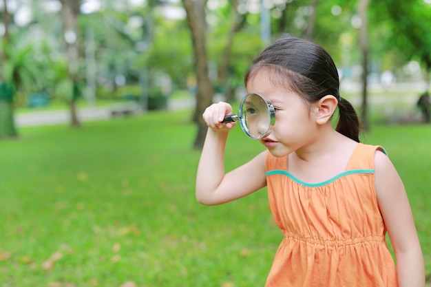 Pequeña muchacha asiática linda del niño que mira a través del vidrio magnifiying encendido la hierba al aire libre.