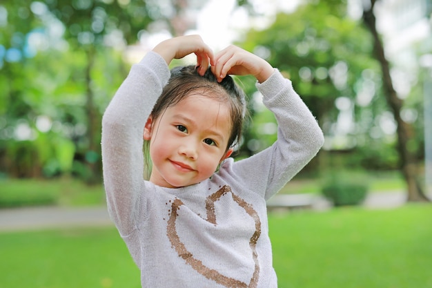 La pequeña muchacha asiática linda sonriente del niño en jardín verde con la fabricación sus manos para la muestra del corazón