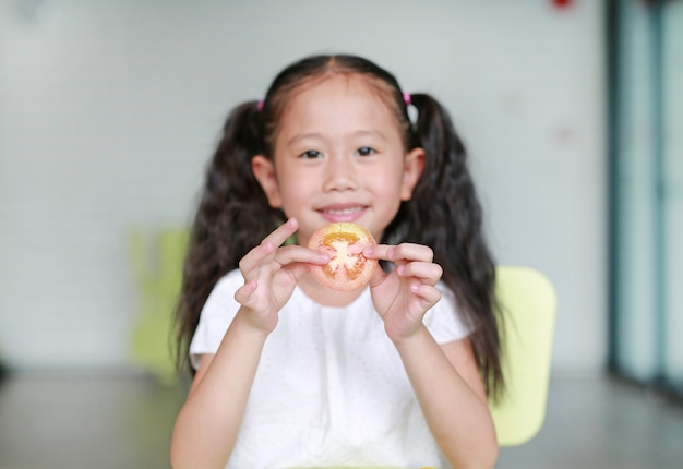 Pequeña muchacha asiática sonriente del niño que sostiene un pedazo de tomate rebanado. Niño comiendo concepto de comida saludable. Centrarse en tomate en manos de los niños.