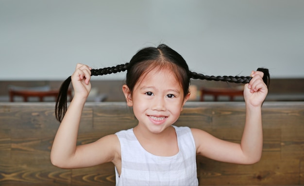 Pequeña muchacha asiática sonriente que sostiene su pelo de las coletas que miente en la tabla de madera con la mirada de la cámara.