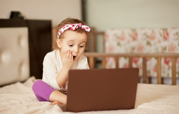 Pequeña muchacha linda que se sienta en la cama y que usa un cuaderno portátil de la tableta digital. Llamadas en línea a amigos o padres.