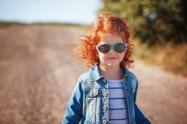 Pequeña muchacha rizada roja linda del retrato en gafas de sol en día soleado