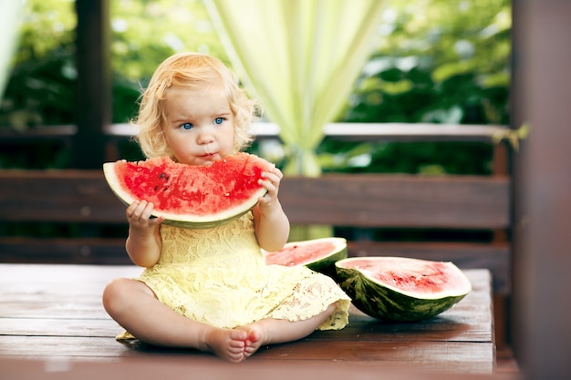 Pequeña muchacha rubia que come una sandía jugosa en el jardín. Los niños comen fruta en la calle. Comida sana para niños. Jardinería para niños pequeños.