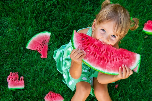 Pequeña muchacha rubia que se sienta en la hierba y que come la rebanada de sandía en verano.