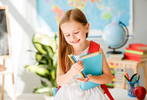 Pequeña muchacha rubia sonriente que sostiene el libro azul y que mira los lápices coloridos en la clase de escuela del sol