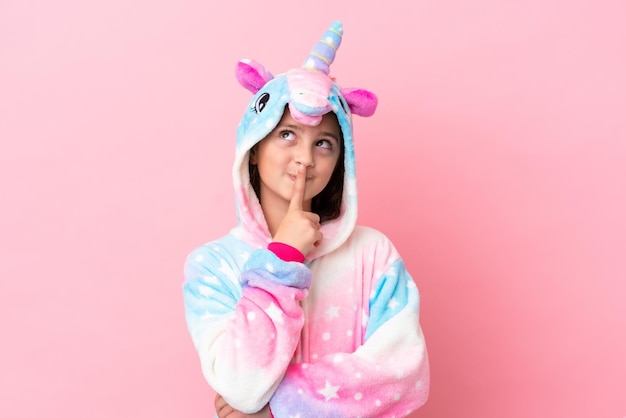 Pequeña mujer caucásica con un pijama de unicornio aislado en un fondo rosado que tiene dudas mientras mira hacia arriba