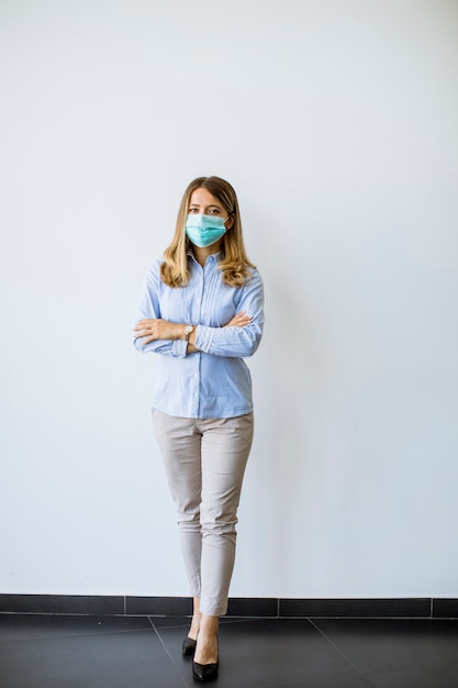 Pequeña mujer de negocios joven con mascarilla protectora se encuentra junto a la pared en la oficina