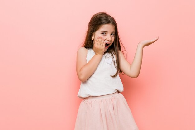 La pequeña mujer que lleva una mirada de princesa tiene espacio de copia en una palma, mantenga la mano sobre la mejilla. Sorprendido y encantado.