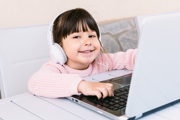 La pequeña niña con auriculares blancos, vistiendo un suéter rosa, sentada frente a la computadora portátil en la terraza, sonriendo. Concepto de educación a distancia en línea, niñez y tecnología.