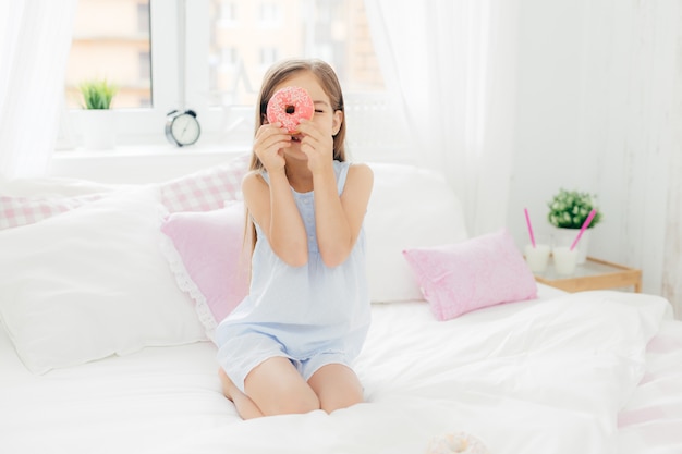 Pequeña niña bonita tiene sabrosa rosquilla dulce, posa en el dormitorio en una cama cómoda