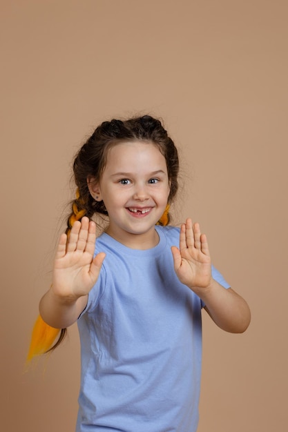 Pequeña niña caucásica fascinada con trenzas kanekalon mostrando las palmas de las manos jugando sonriendo con un diente faltante mirando la cámara en un fondo beige con una camiseta azul