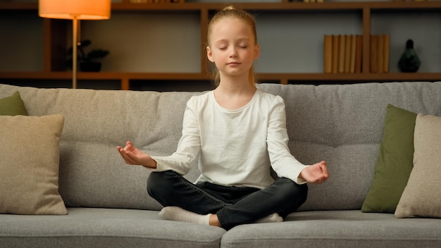 Pequeña niña caucásica linda niña rubia meditar en el sofá en casa calma relajarse solo en posición de loto