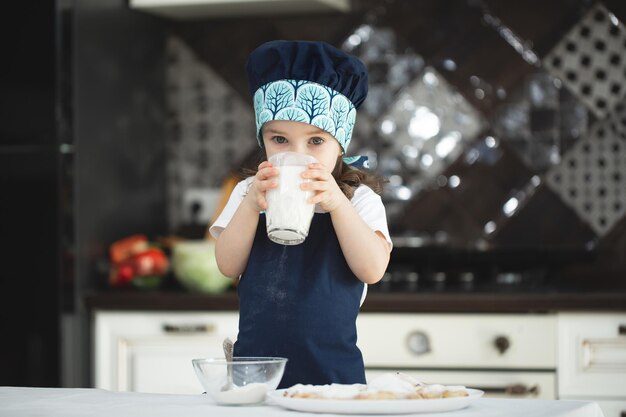 La pequeña niña en la cocina con un delantal y un gorro de cocinero está bebiendo leche de un vaso de vidrio.
