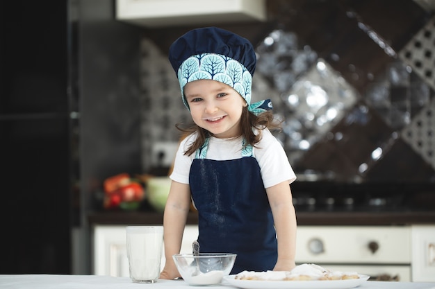 La pequeña niña en la cocina con un delantal y gorro de cocinero rocía galletas con azúcar en polvo