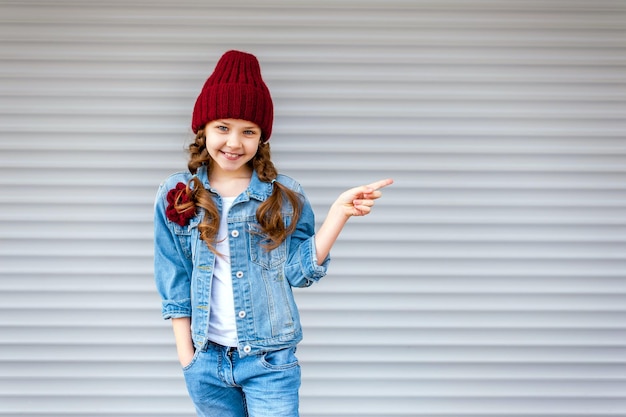Una pequeña niña elegante y de moda se pone de pie con un traje de mezclilla y señala con el dedo