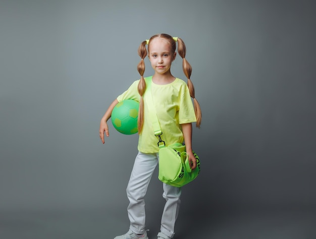 Una pequeña niña con una espada verde en la mano sonríe a la cámara aislada en un fondo gris un pequeño atleta con una pelota de deportes para niños