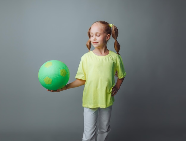 Una pequeña niña con una espada verde en la mano sonríe a la cámara aislada en un fondo gris un pequeño atleta con una pelota de deportes para niños