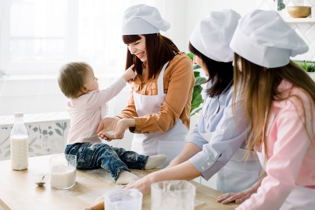 La pequeña niña está sentada en la mesa de madera en la cocina y se divierte con el azúcar. La abuela y sus hijas están horneando galletas. Mujeres felices en delantales blancos para hornear juntos. Día de la Madre