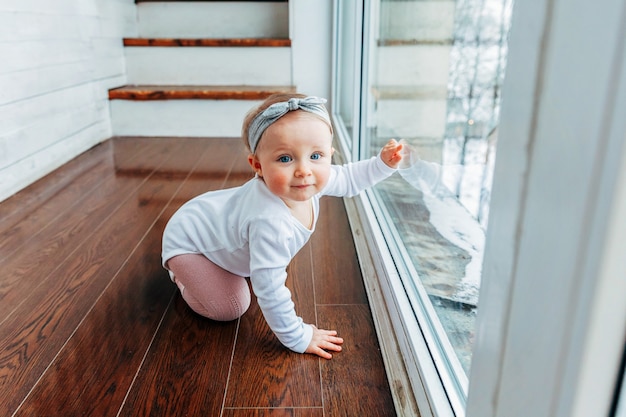 Pequeña niña gateando en el suelo en la sala de estar con luz brillante junto a la ventana sonriendo