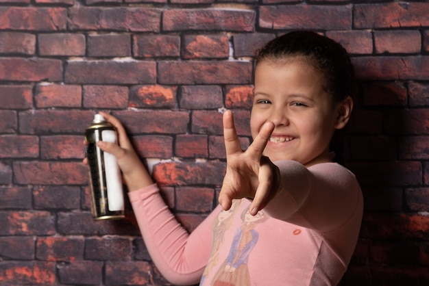 Pequeña niña haciendo poses con un spray frente a un viejo telón de fondo de pared de ladrillo, fondo oscuro, enfoque selectivo.