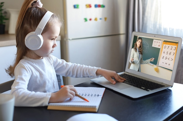 La pequeña niña inteligente en edad preescolar en auriculares mira la lección en línea y se comunica con el maestro en casa, el niño pequeño en auriculares estudia en Internet usando una conexión inalámbrica de computadora portátil.