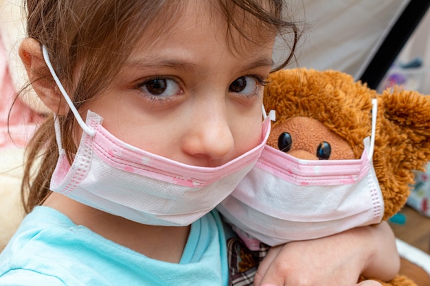 Foto pequeña niña jugando oso de juguete en máscara protectora médica durante el aislamiento de cuarentena