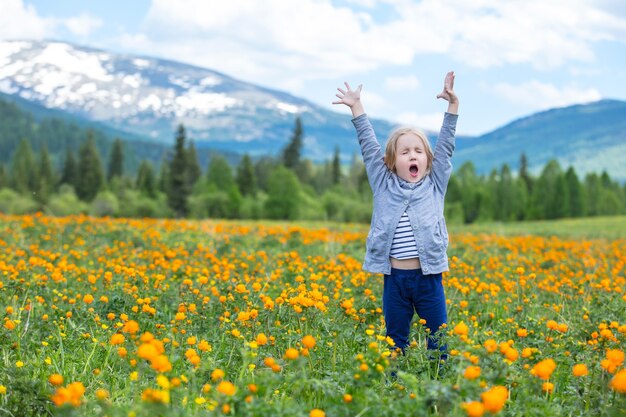 La pequeña niña linda es hermosa y feliz, sonriendo en verano en el prado contra las montañas con nieve