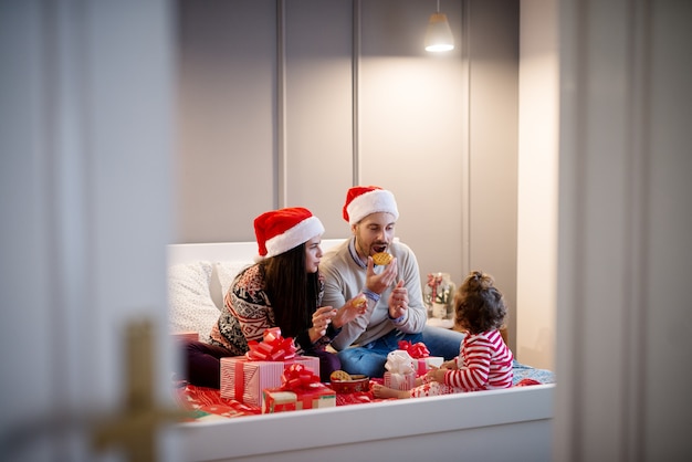 Pequeña niña pequeña rizada que mira en padres mientras que come y juega con las galletas en la cama para las vacaciones de Navidad.