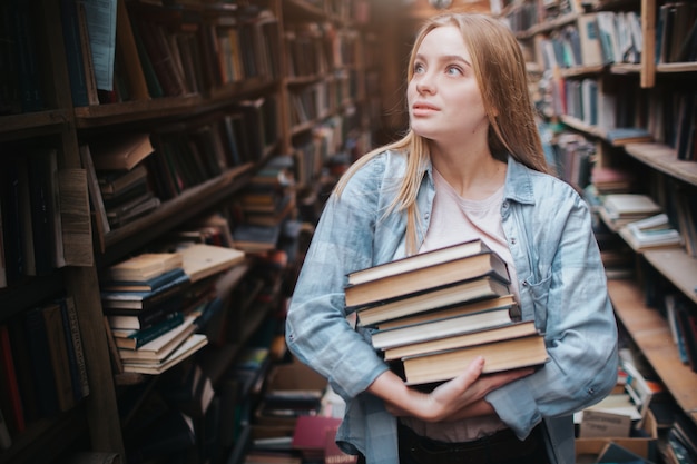 Una pequeña niña de pie en una gran librería vieja y sosteniendo muchos libros en sus manos. Ella está buscando otro libro para llevar con ella. La joven belleza se ve soñadora.