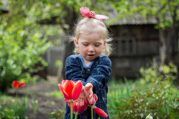 La pequeña niña rubia camina en el jardín cerca de tulipanes rojos florecientes rojos.