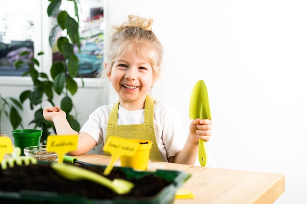 Una pequeña niña rubia con un delantal se dedica a plantar semillas para plántulas, sonriendo, el concepto de jardinería infantil.