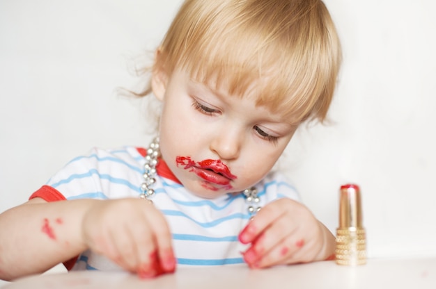 La pequeña niña rubia divertida compone los labios con el lápiz labial rojo de la mamá. Aprendiendo a ser mujer.