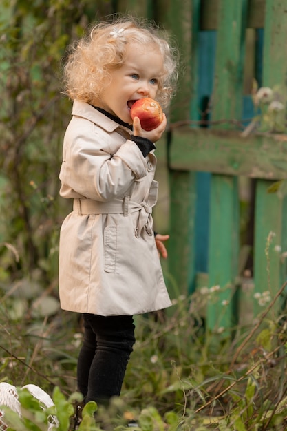 Pequeña niña rubia de pelo rizado con ojos azules vestida con un impermeable blanco come una manzana al aire libre