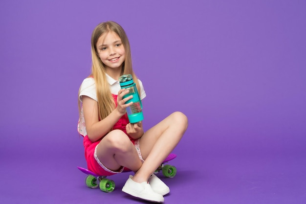 La pequeña niña se sienta en patineta con botella sobre fondo violeta. Sonrisa de niño patinador con longboard. Niño patineta en mono rosa. Deporte, actividad y energía. Estilo de vida infantil y juegos activos.