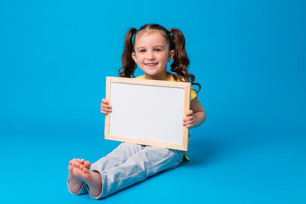 La pequeña niña sonríe y sostiene un tablero de dibujo vacío en azul