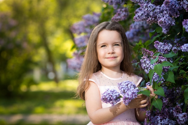 Una pequeña niña de sonrisa está en los arbustos de lilas en el jardín al atardecer, parque floreciente de primavera.