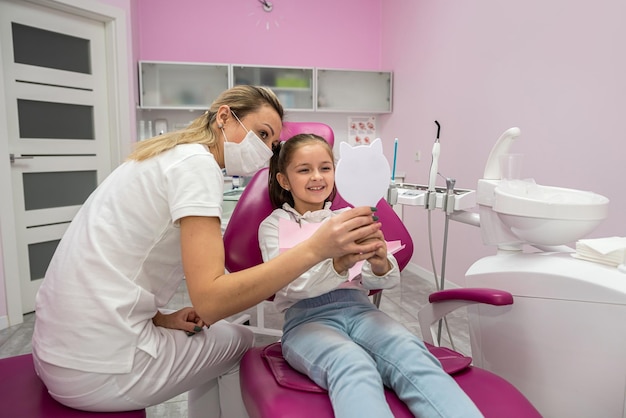 Pequena paciente na cadeira dentária olha para os dentes depois do tratamento