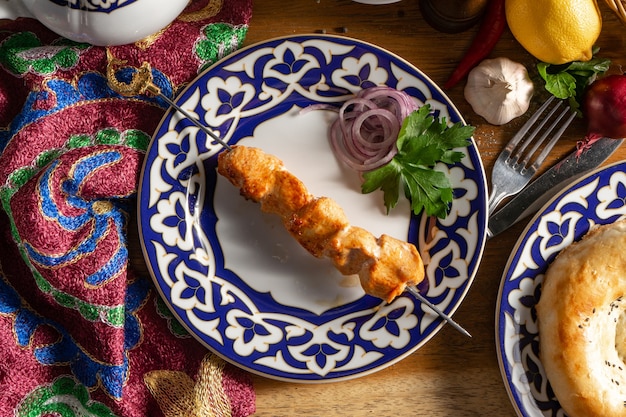 Una pequeña porción de shish kebab de pollo en un pincho con cebolla morada y perejil en un plato con un patrón nacional de Uzbekistán