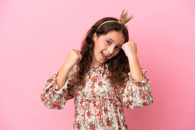 Pequena princesa caucasiana com coroa isolada em fundo rosa comemorando uma vitória