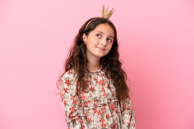 Pequena princesa caucasiana com coroa isolada em fundo rosa pensando em uma ideia enquanto olha para cima